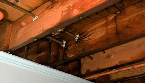 Exposed overhead wood framing and pipes above an interior ceiling opening